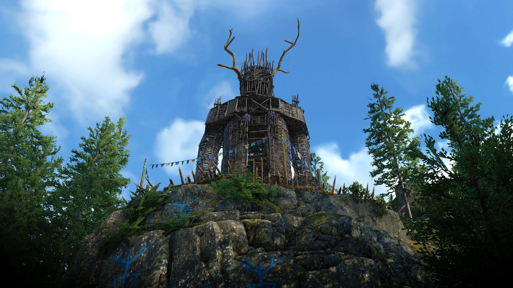 A screenshot showing a wicker man atop a rocky outcrop, with wooden stakes set up around it and tattered blue, white, and cream flags. Large trees stand alongside it, and the sides of the rocky outcrop are painted with strange blue paganistic markings.