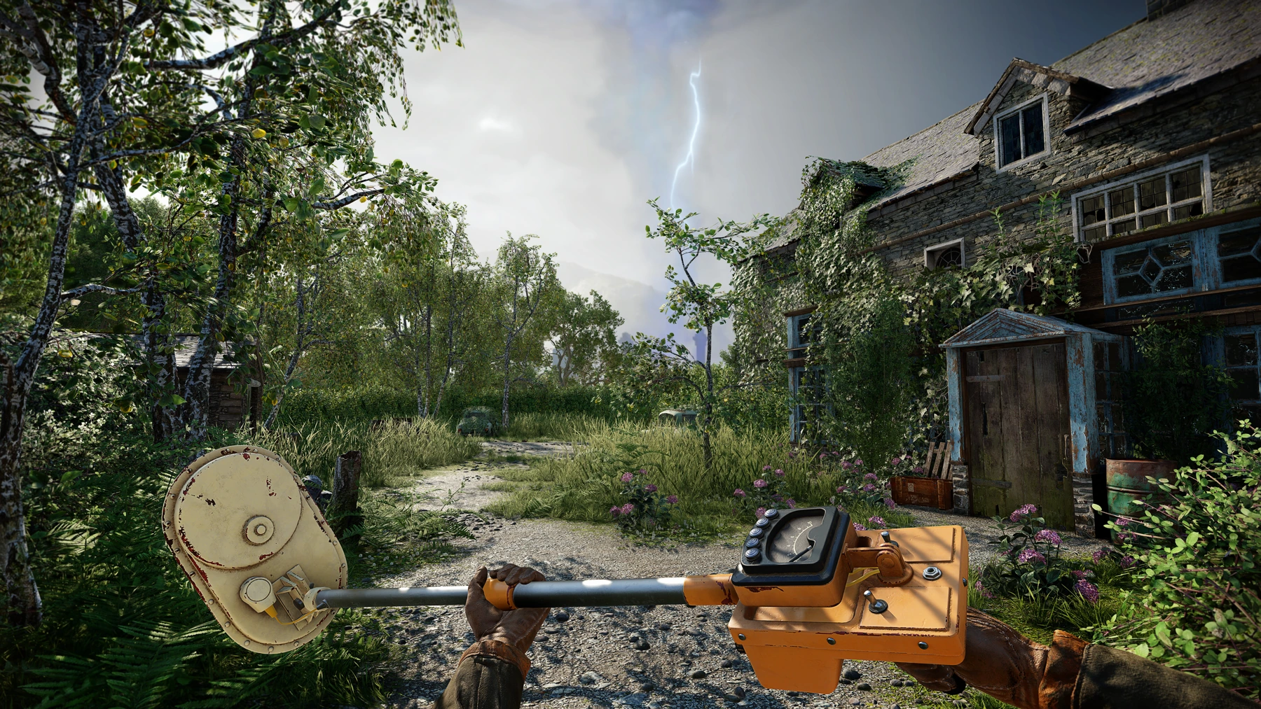 A screenshot with a first-person view of a character holding an orange metal detector, looking out at an overgrown country mansion and a garden full of weeds and overgrown plants and trees. A gravel path leads through the garden, with ruined cars and the silhouette of a nuclear power plant in the background. Strange blue light is emanating from the power plant, with a bolt of lightning striking it.